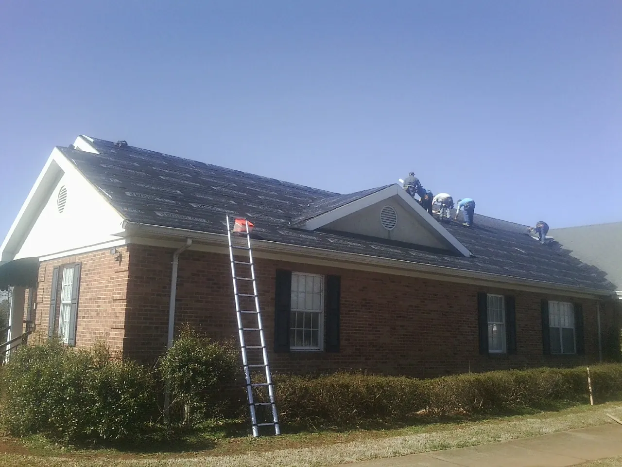 Crew tearing shingles off a brick ranch roof with a ladder set — Emergency Roof Repair work by our crew in Pleasant Lake